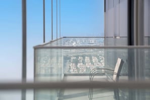 Une vue depuis une fenêtre, avec un ciel dégagé, un balcon avec une chaise, et un paysage urbain lointain.