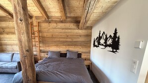 A bedroom with a wooden ceiling, a bed with grey bedding, a grey sofa, and a wall with a black forest mural. - Le mazot - large studio with separate shower room (Lens)
