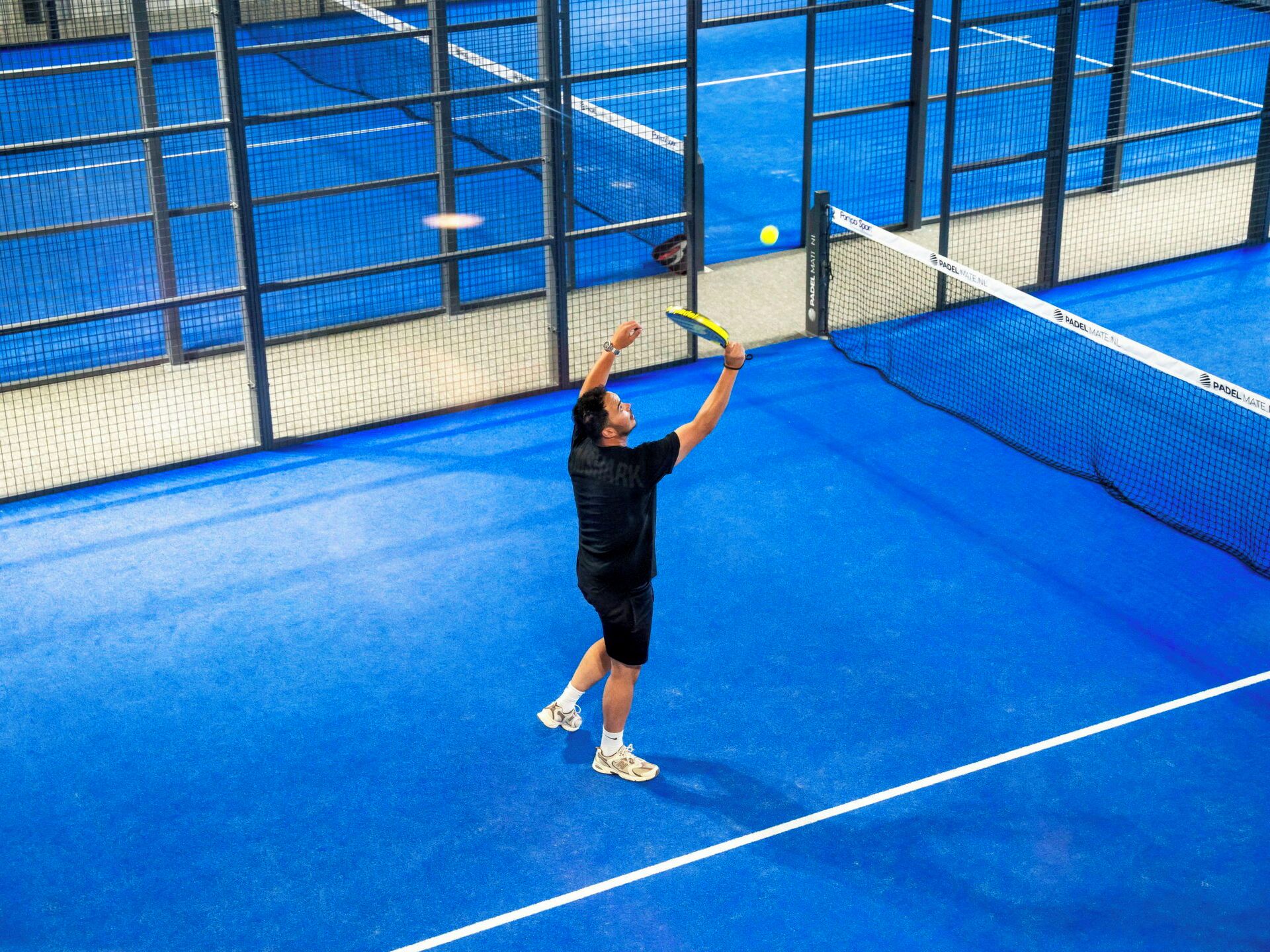 Een tennisser in volle actie op een blauwe indoorbaan, met een net en een bal in het spel.