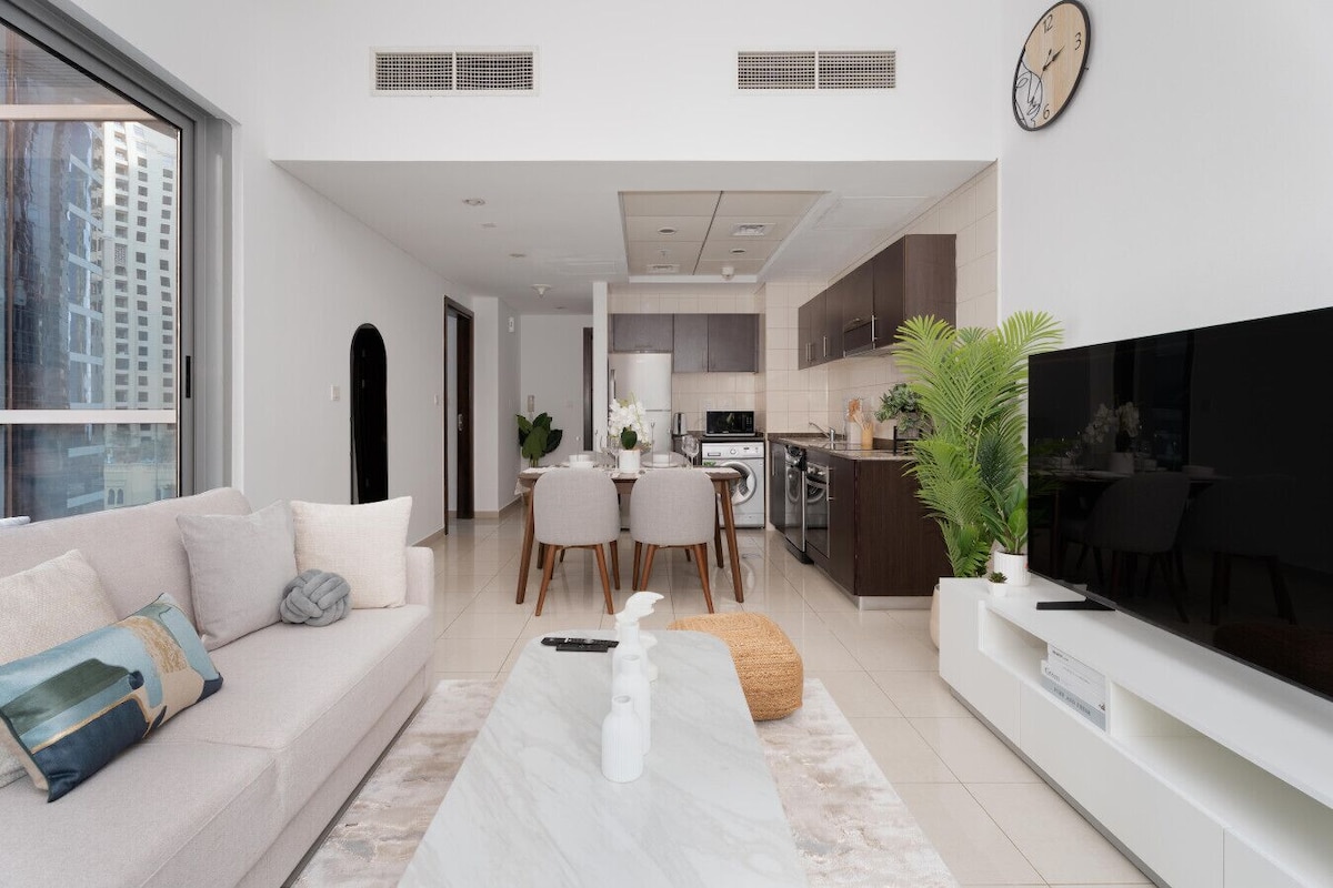 Ein modernes Wohnzimmer mit einer Couch, einem Couchtisch und einem Fernseher. Im Hintergrund befindet sich ein Essbereich mit Stühlen und eine Küche.