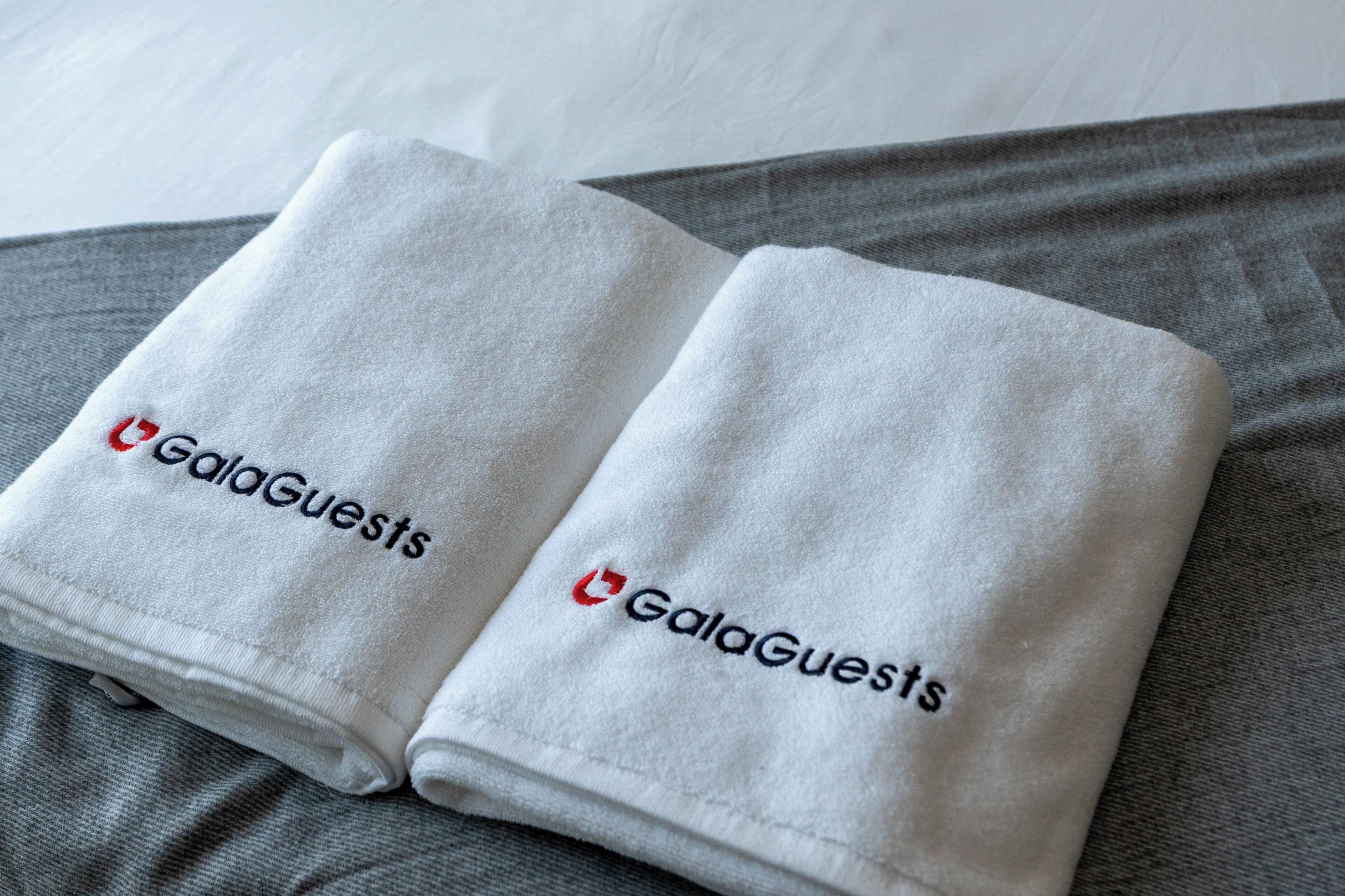 Two folded white towels with the GalaGuests logo on a grey fabric surface.
