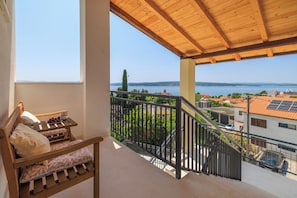 Ein Balkon mit Holzboden, ein Bett mit Kissen und ein kleiner Tisch mit Früchten, mit Blick auf ein Gewässer und Gebäude.
