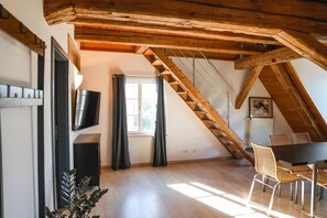 Une pièce avec un plafond en bois, un téléviseur fixé au mur, une fenêtre avec des rideaux, une table à manger avec des chaises et un escalier.