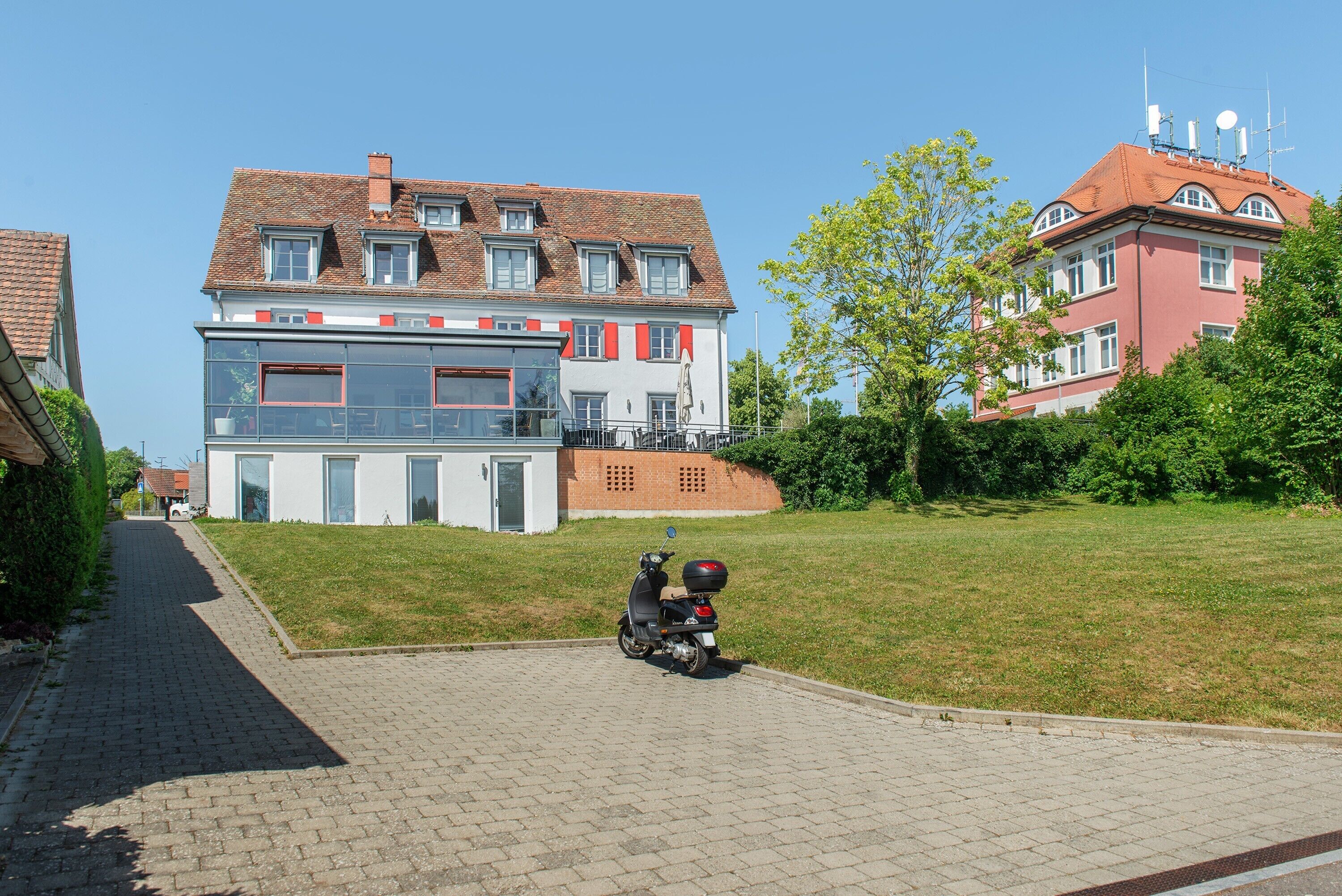 Une allée pavée mène à un bâtiment moderne à deux étages, doté d’un toit rouge, de grandes fenêtres et d’une porte de garage bien visible.