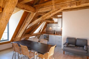 Un loft moderne avec des poutres apparentes, un coin repas avec des chaises en osier, un coin cuisine et un canapé gris.