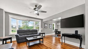 Ein Wohnzimmer mit grauem Sofa, Holzboden, Deckenventilator und Wand-Fernseher.