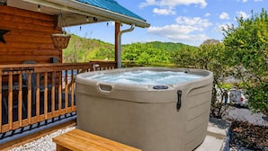 Ein Whirlpool vor einer Holzhütte mit blauem Dach, umgeben von viel Grün und blauem Himmel.