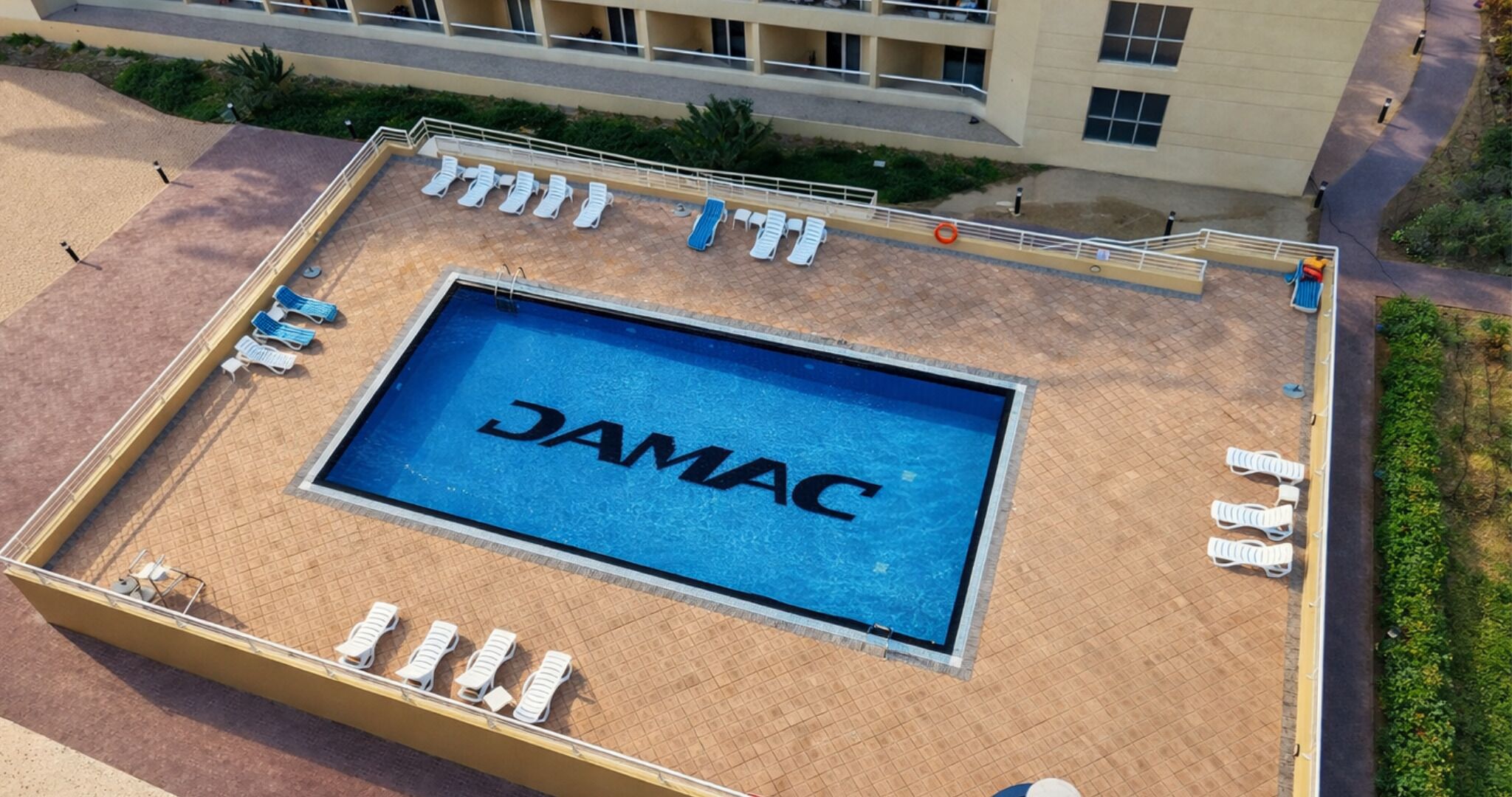 Vue aérienne d’un espace piscine avec un panneau indiquant « JAMAC » et plusieurs chaises longues disposées autour de la piscine.