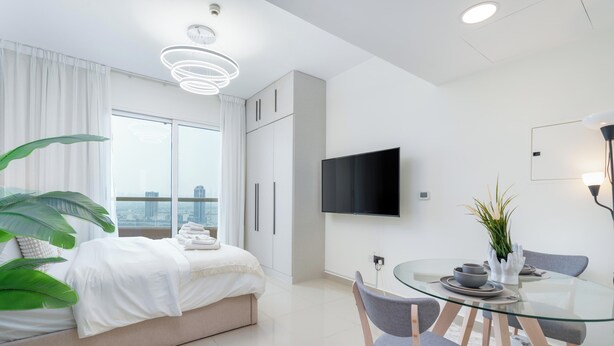 A modern hotel room with a bed, a dining area with a glass table, and a TV on the wall. - Gala Guests - Lakeside Tower Living (Dubai)