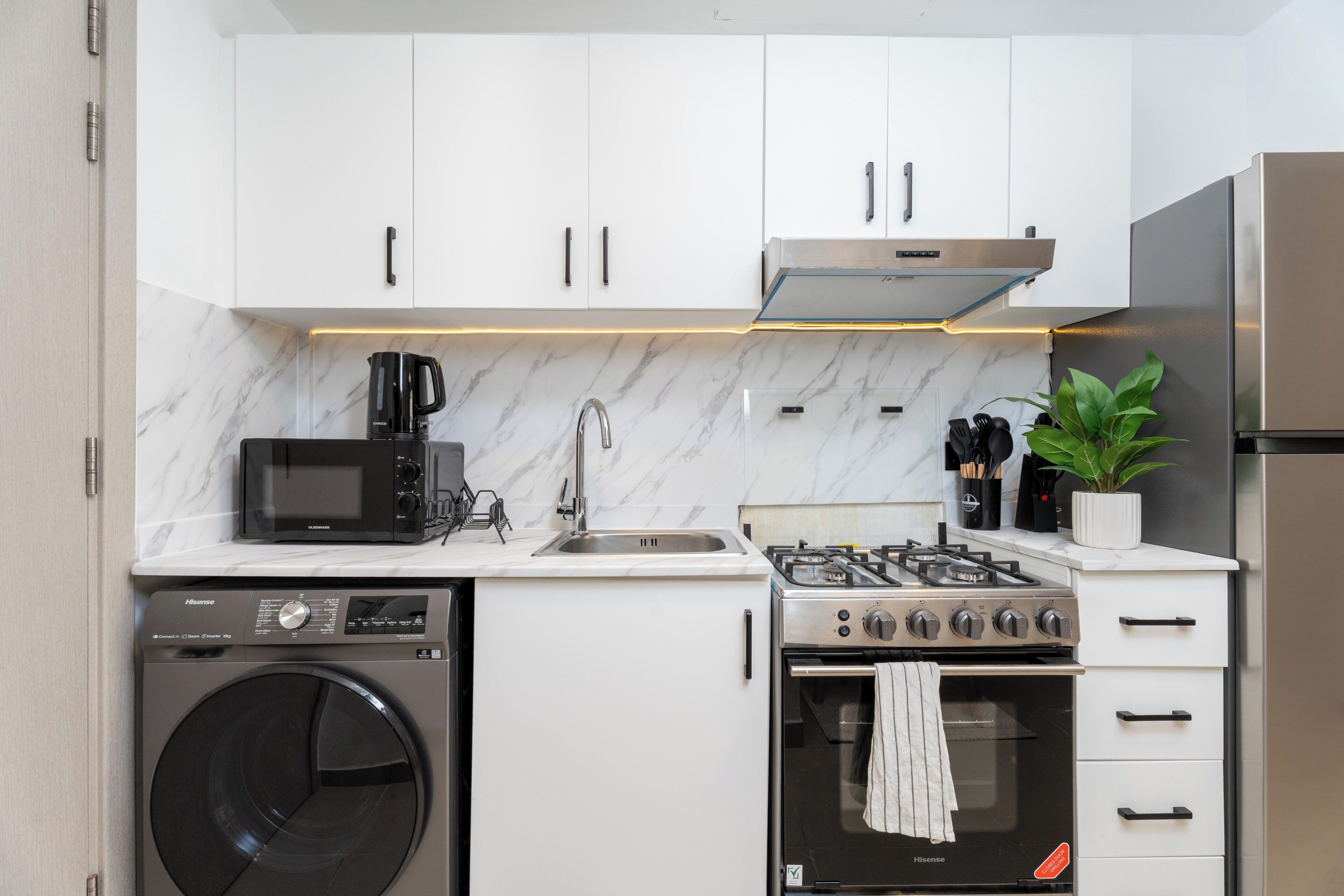 Cocina compacta con armarios blancos, microondas integrado, estufa a gas, lavadora y refrigerador de acero inoxidable.