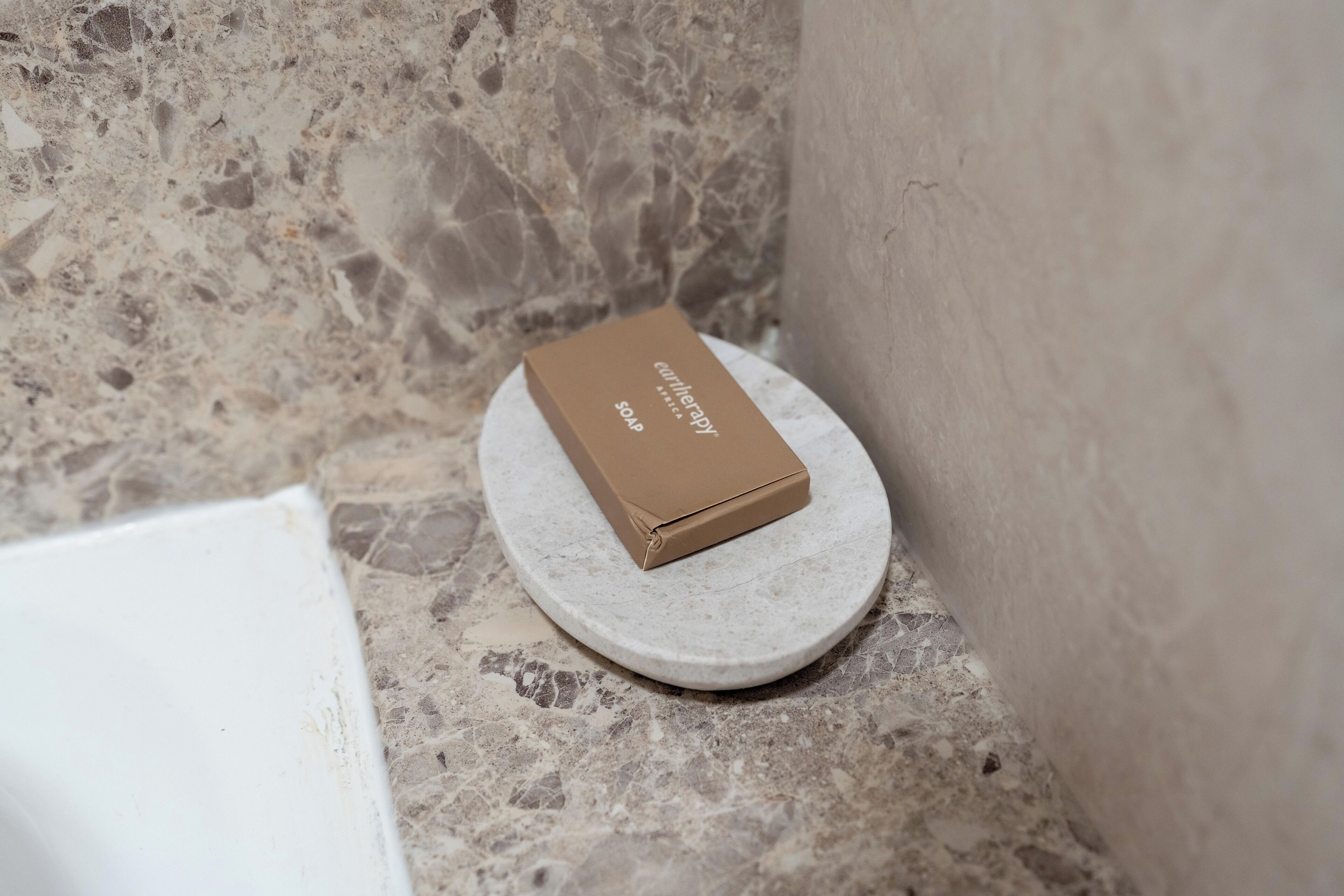 A small, round, white stone pedestal with a brown box labeled "curiltherapy" on top, placed in a corner with a textured stone wall and a plain wall in the background.