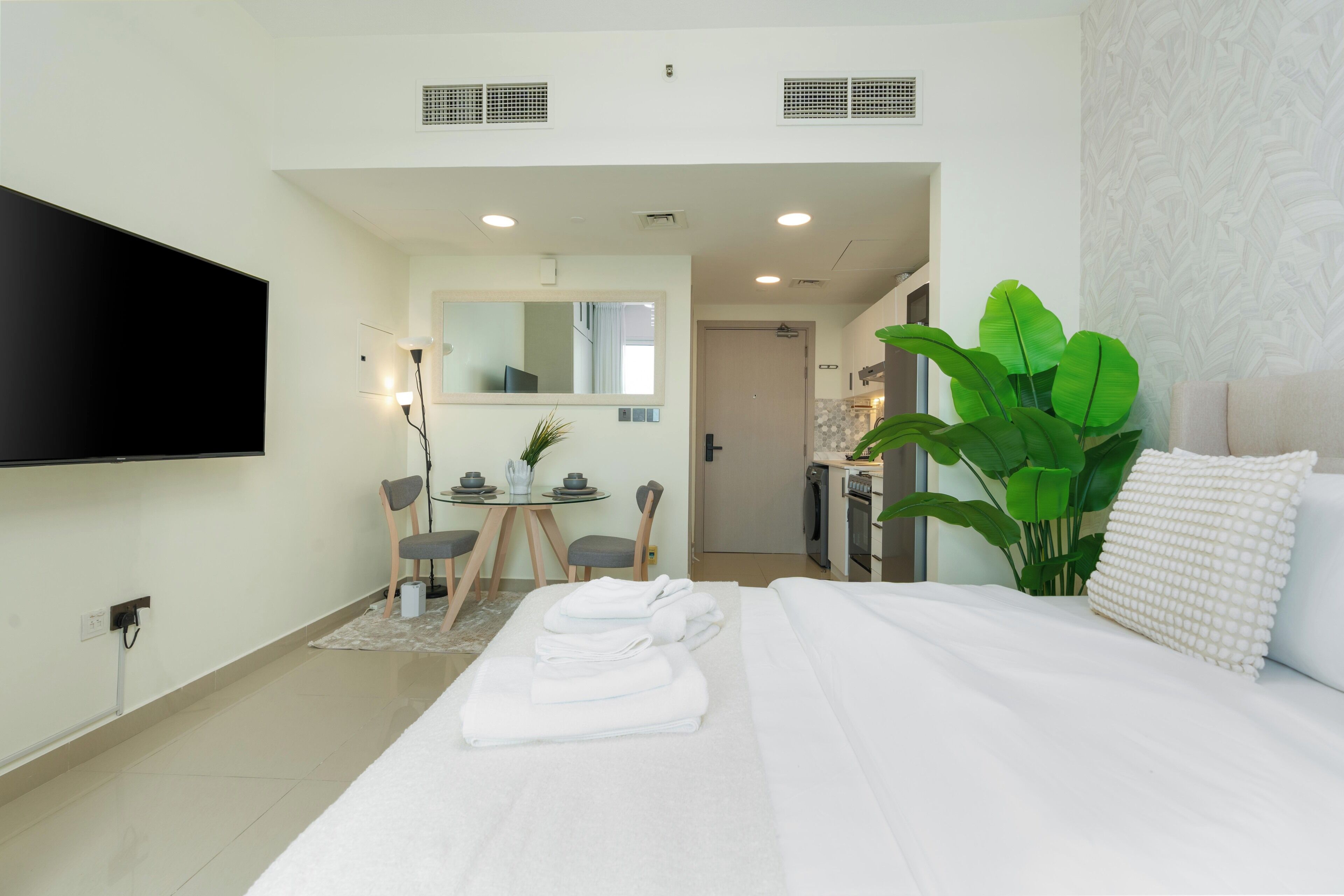 Quarto de hotel com cama, televisão, secretária com cadeiras e uma planta grande.