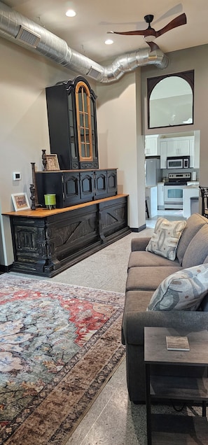 A living room with a sofa, a coffee table, a fireplace, a kitchen in the background, and a ceiling fan.