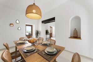 Ein Esszimmer mit einem Holztisch, gedeckt für vier Personen, umgeben von Korbstühlen, einer Hängelampe und einer Pflanze im Hintergrund.