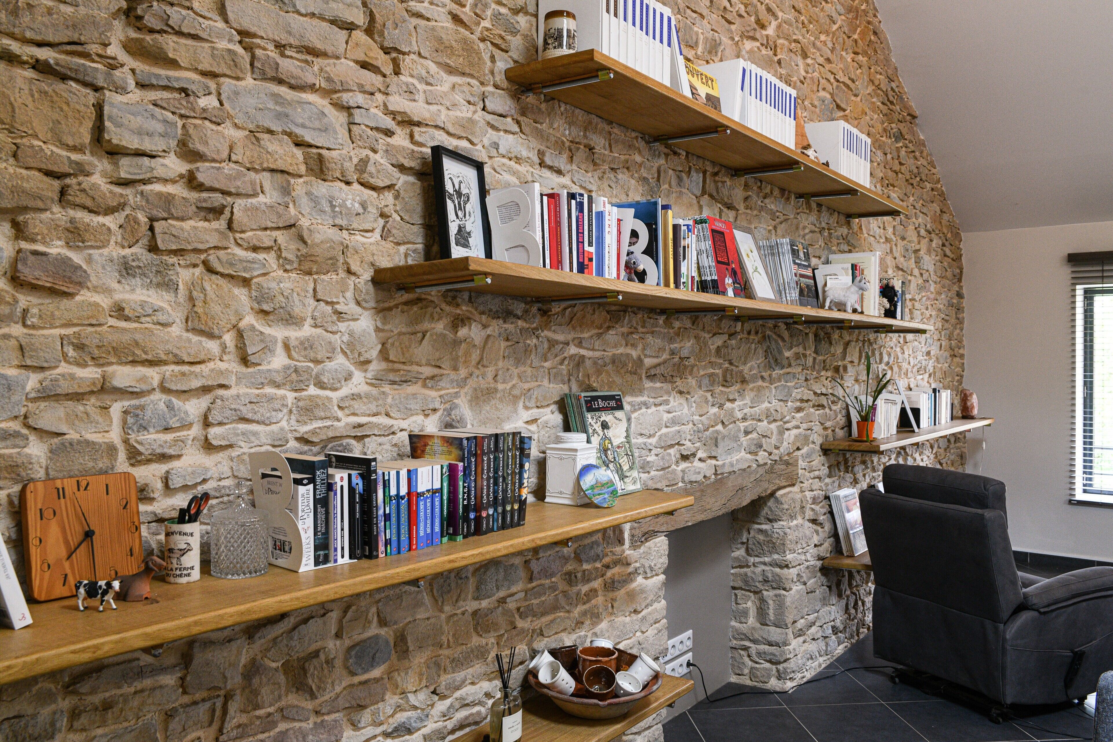 Un mur en pierre avec des étagères où sont posés des livres et des objets décoratifs, une horloge et une cheminée.