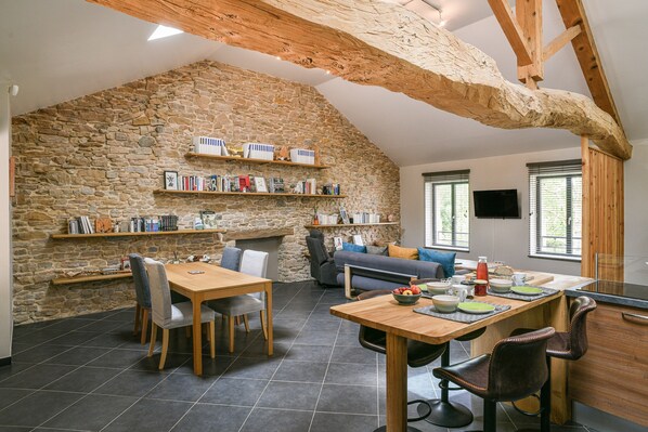 Un loft spacieux avec un mur en pierre, des poutres apparentes, un coin repas et un espace salon comprenant un canapé et des fauteuils.