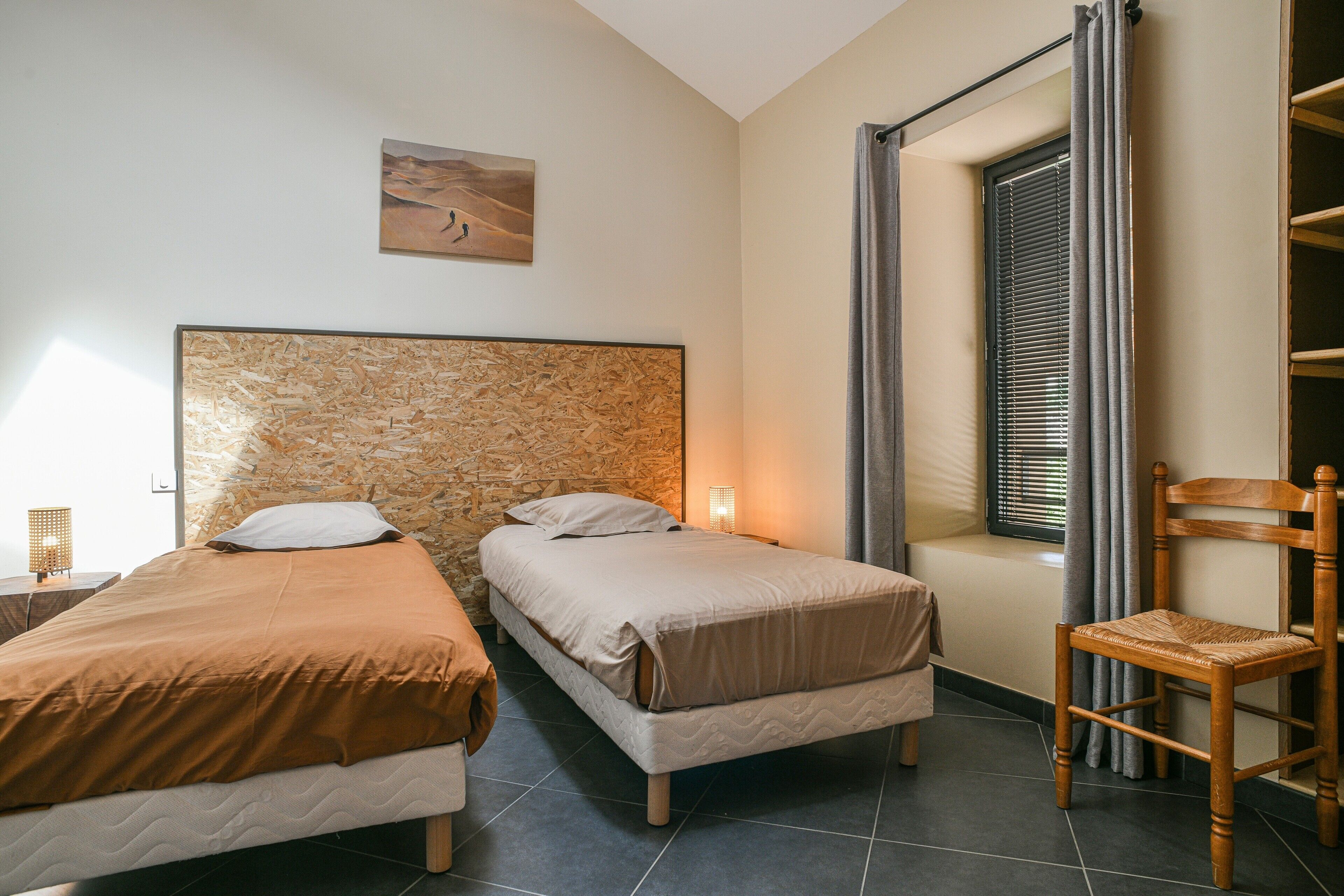 A room with two beds, a wooden headboard, a chair, and a window with curtains.
