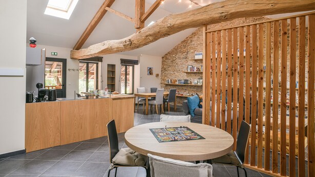 A cozy interior with a wooden dining table, chairs, a kitchen area, and a stone wall bookshelf. - Gîte "La Grange d'Émile" with Private Garden and Wi-Fi (Chozeau)