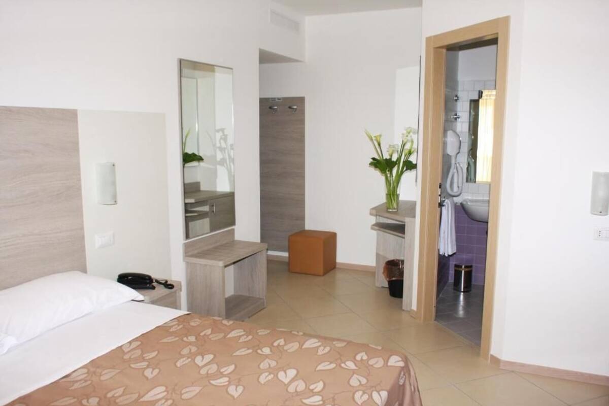 Quarto de hotel com cama, banheiro com chuveiro, penteadeira com espelho e um banquinho pequeno.