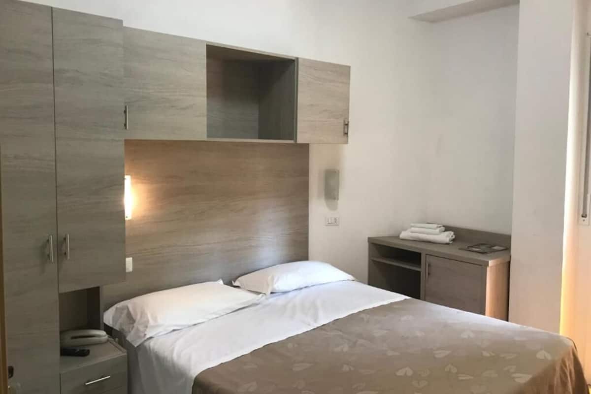 Um quarto com cama, guarda-roupa, mesa de cabeceira e apoio para luminária.