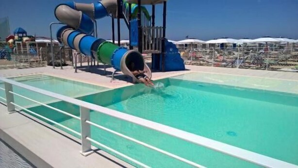 Una piscina con tobogán, un parque infantil y sombrillas al fondo.