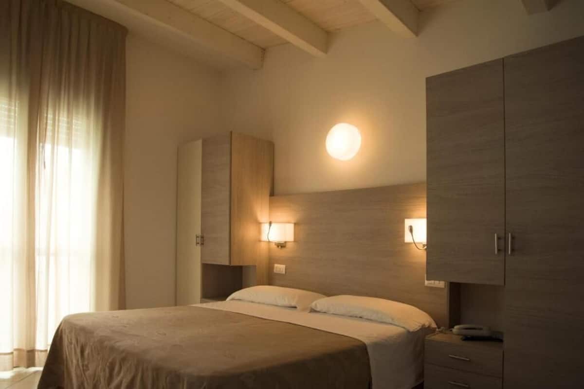 Quarto de hotel com cama, abajures, guarda-roupa e uma luminária redonda na parede.