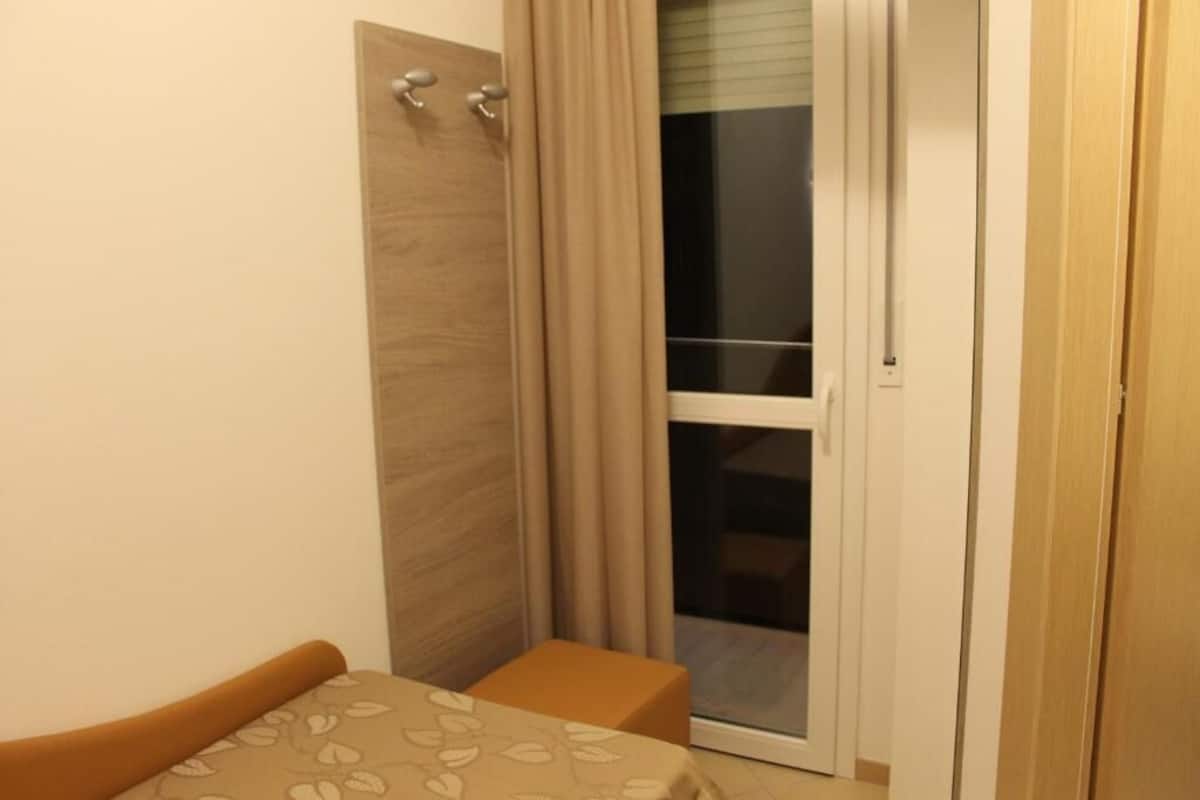 Um quarto pequeno com uma cama, uma porta de madeira, uma porta de correr de vidro e uma mesinha.