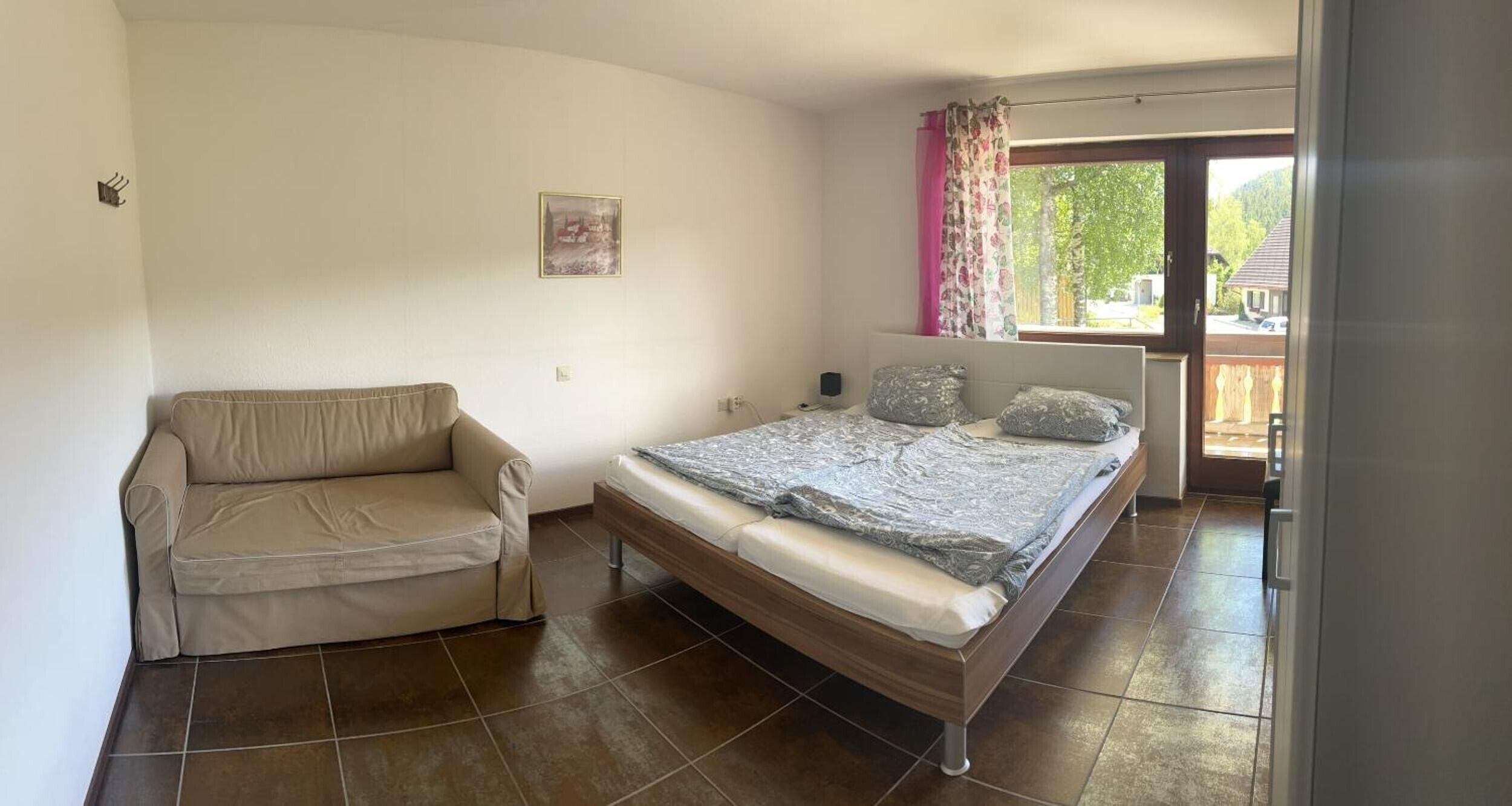 A bedroom with a bed, a sofa, a window with curtains, and a door leading to an outdoor area.