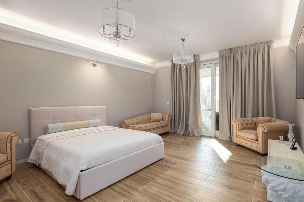 Un dormitorio con una cama grande, dos sillones, una mesa de centro de vidrio, una araña y un cerramiento de vidrio corredizo que da a un balcón.