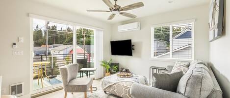 A modern living room with a ceiling fan, large windows, a flat-screen TV, a sofa, and a dining area with a table and chairs.