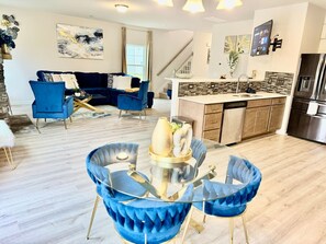Una sala de estar moderna con un sofá azul, una mesa de centro de vidrio y una zona de cocina con un refrigerador de acero inoxidable.
