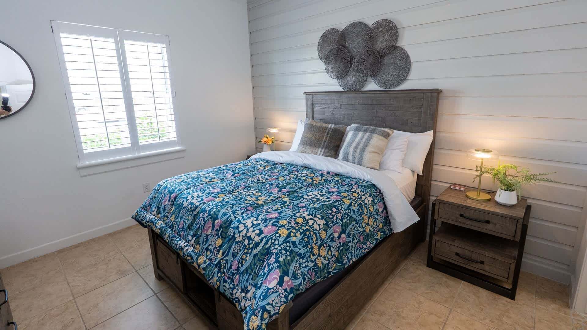 A bedroom with a bed, a nightstand, a wall-mounted decorative piece, and a window with blinds.