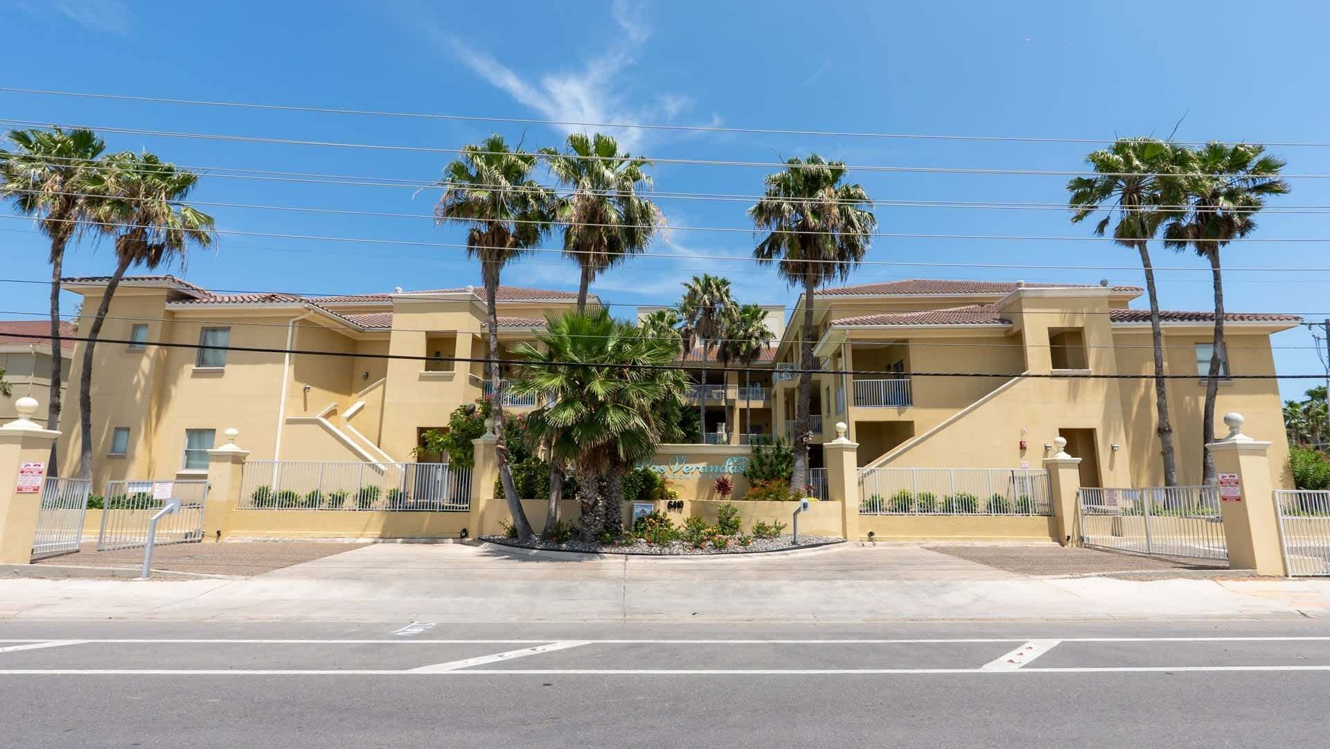 A multi-story hotel with a beige facade, palm trees, and a gated entrance.