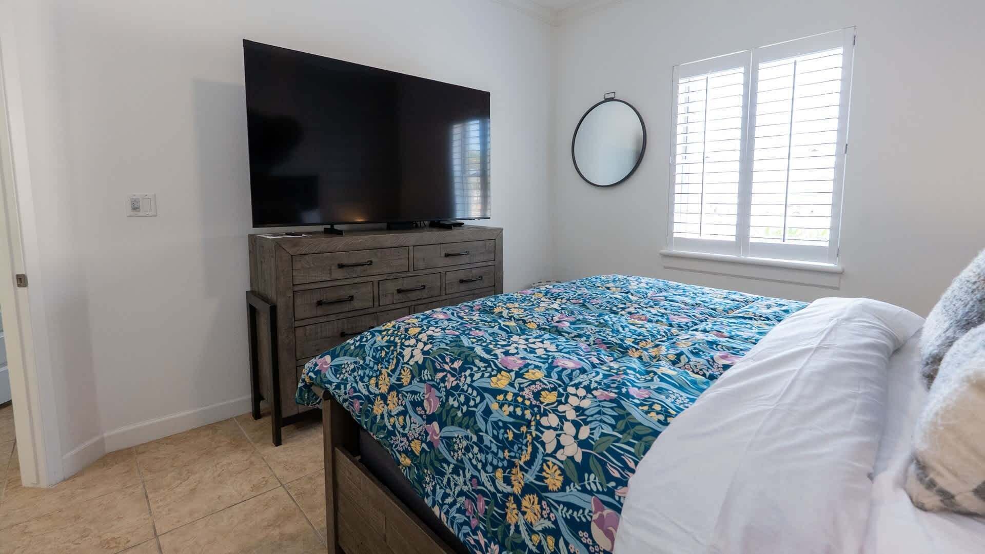 Een slaapkamer met een bed, een dressoir, een televisie, een raam met zonwering en een spiegel aan de muur.