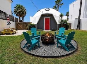 Quintal com lareira, quatro cadeiras Adirondack azuis, uma porta vermelha e um edifício branco com telhado em forma de cúpula.