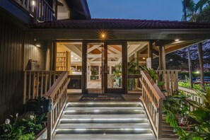 A hotel entrance with a wooden staircase, a covered porch, and a well-lit interior visible through the glass doors. - Remodeled Ground Floor 2BR Steps to Pool & Hot Tub -Papakea K105 (Lahaina)