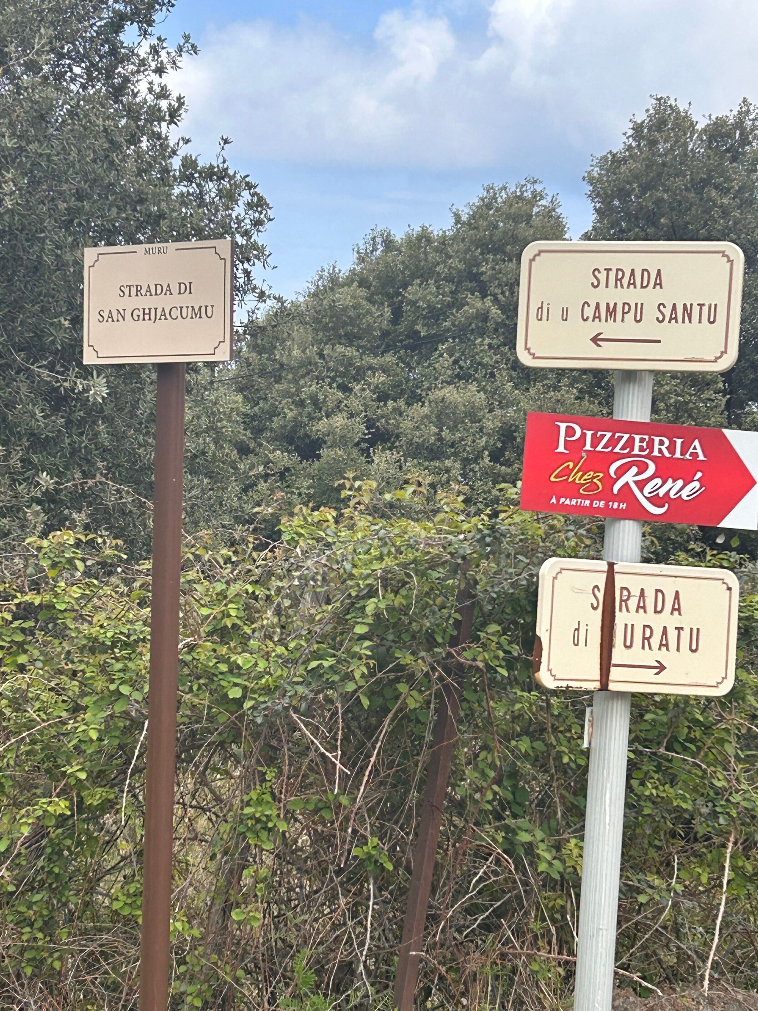 Skilte der viser vej til Strada di San Ghjacumu, Strada di u Campu Santu og Pizzeria Chez Rene.