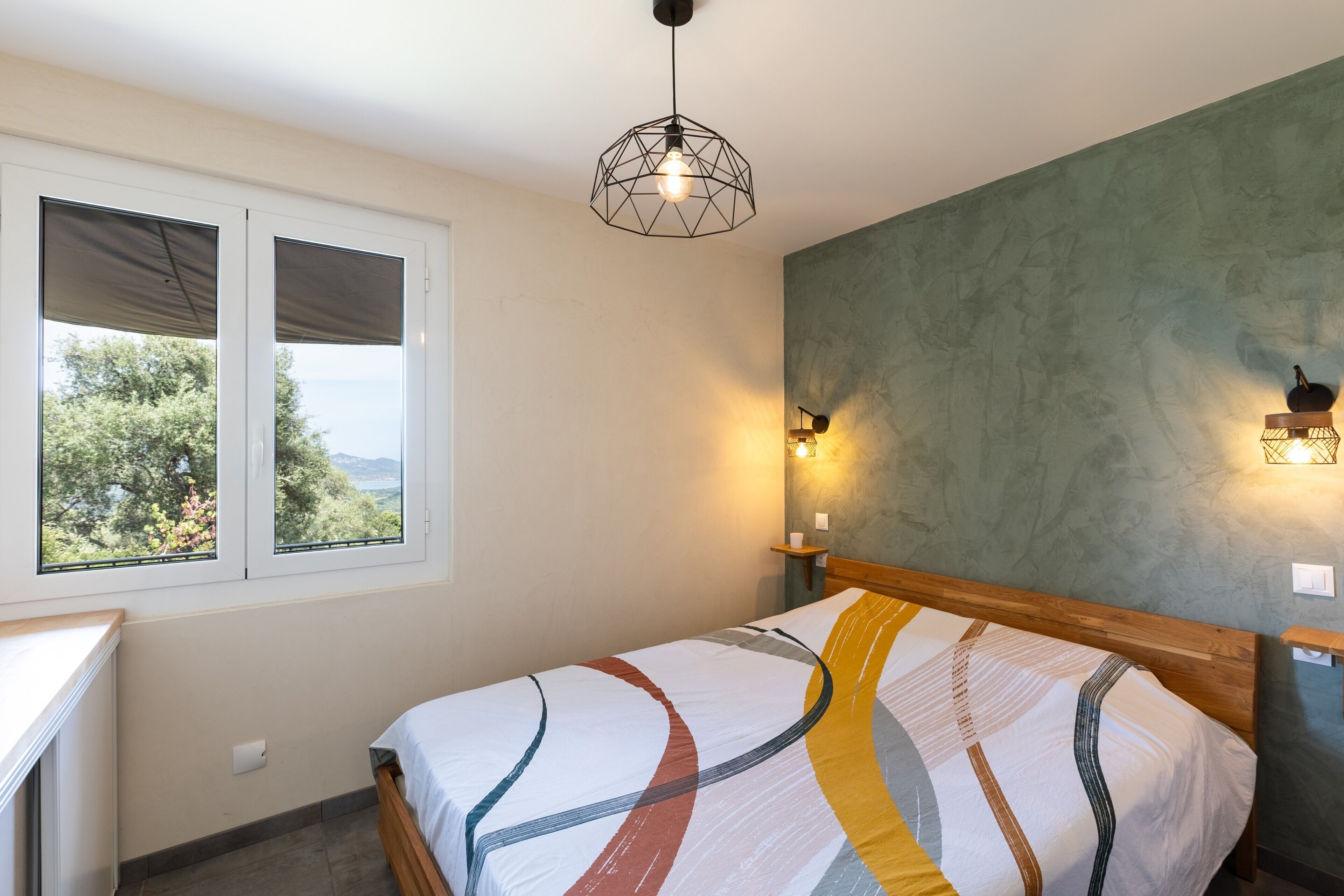 Une chambre à coucher avec un lit, une fenêtre avec des stores, un luminaire fixé au mur et un autre suspendu au plafond.