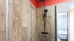 Doccia con soffione a mano, pannello di controllo a parete e porta in vetro che dà accesso a una camera da letto con un letto e una lampada a sospensione.