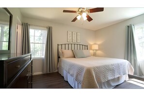 Ein Schlafzimmer mit Bett, Schminktisch, Deckenventilator und einem Fenster mit Vorhängen.