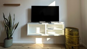 Ett modernt vardagsrum med en platt-TV placerad på en vit TV-bänk, en krukväxt och en stor tunna.