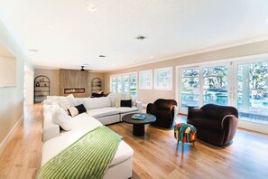 A spacious living room with a white sectional sofa, a round coffee table, and two leather armchairs. Large windows provide a view of a swimming pool. - Indian Rocks Escape Luxury Beach Villa Pickleball, Pool & Game Room (Largo)