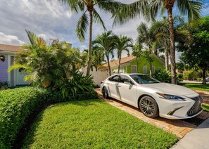 A white car parked on a paved driveway with a well-maintained lawn and palm trees. - Coco Plum Cottage a Charming Retreat Htd Pool and BBQ (West Palm Beach)