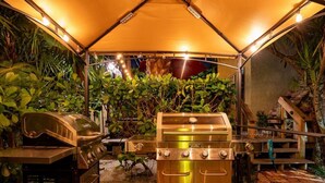 Eine Terrasse mit einem Edelstahlgrill, einem Barbecue-Smoker und einem Sonnenschirm, der Schatten spendet.