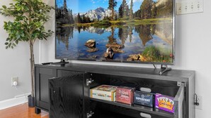 A flat-screen TV on a wooden TV stand with a mountain landscape reflection, a potted plant, and snack boxes. - 3000sqft 5B3.5B Retreat Near Lenovo Center - Upscale 3 King Beds (Cary)