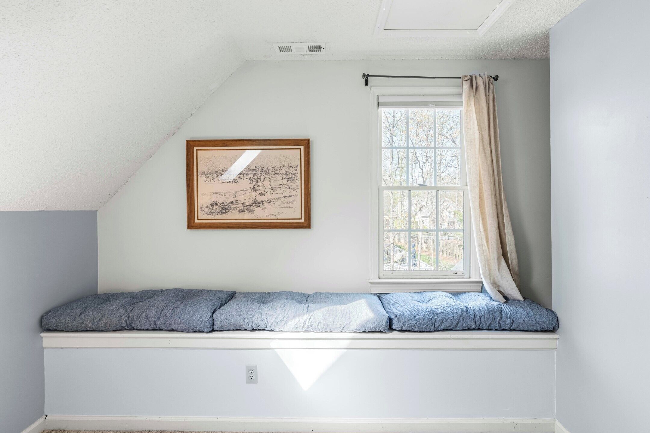 A room with a built-in bench, a window with curtains, a framed picture on the wall, and a ceiling vent.