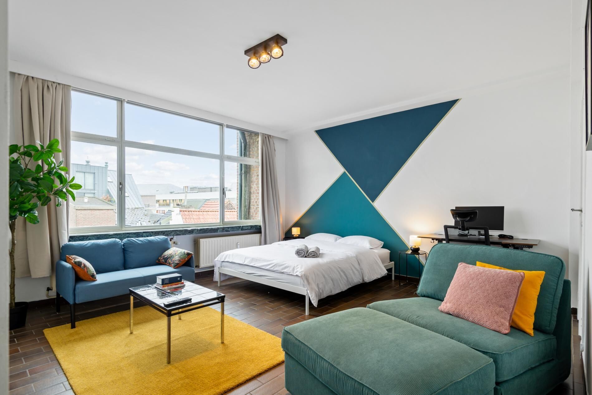 Bright open bedroom and lounge area with a large window overlooking the city. The space combines a comfortable bed, seating area, and workspace, creating a relaxed environment for both rest and quiet moments indoors.