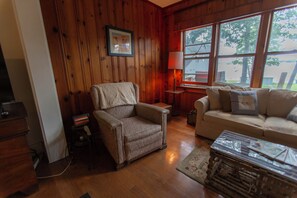 A cozy living room with wooden walls, a comfortable armchair, a sofa, and a lamp. - 4-bedroom Lakefront Retro Cottage on Lake George in Fremont, IN (Fremont)