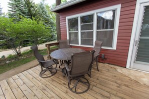 A wooden deck with a round table and four chairs, a window, and a door with a view of trees.