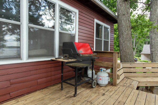 Una terraza de madera con una parrilla y tanque de propano, un banco de madera, y una casa con revestimiento rojo y ventanas blancas.